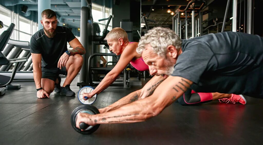 Elderly couple performing an ab rollout workout with a trainer looking on