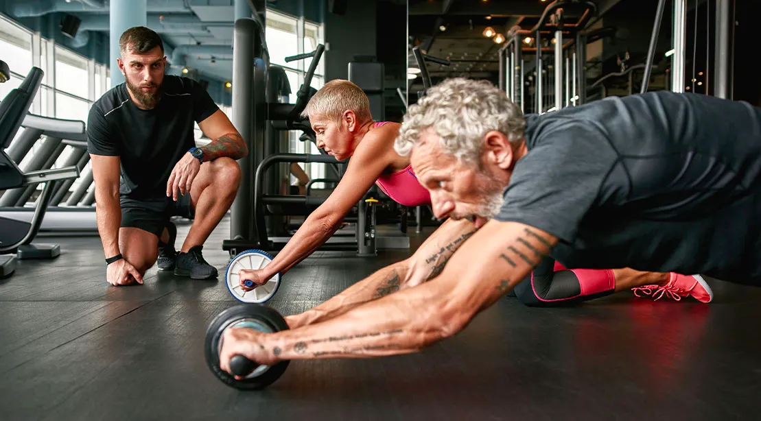 Elderly couple performing an ab rollout workout with a trainer looking on
